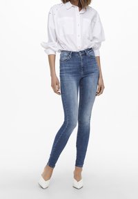 Höga, figursydda blå jeans med en urtvättad textur, kombinerat med en vit skjorta med knappar, som har puffärmar och fickdetaljer.