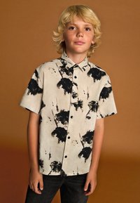 Chemise à manches courtes à boutons en beige clair avec motif abstrait éclaboussé noir. Tissu en coton, col classique et boutons noirs.