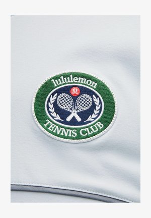 Tissu blanc avec un logo circulaire brodé en vert et bleu marine, texte "lululemon", deux raquettes croisées, et "TENNIS CLUB" en dessous.