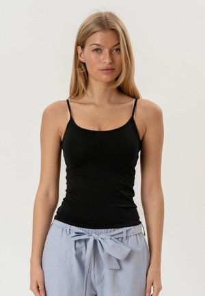 Femeie tânără purtând un top negru cu bretele subțiri și pantaloni drepți cu talie înaltă, albastru deschis cu dungi, având o fundă mare legată în talie.