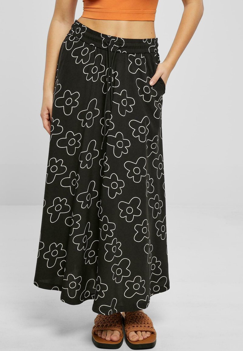 Black maxi skirt with white flower patterns, elastic waistband, and side pockets, worn with brown, woven, flat sandals.