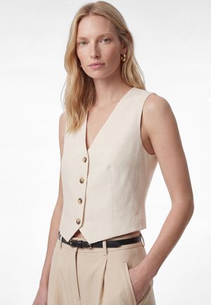 Blonde Frau, die eine ärmellose beige, geknöpfte Weste und dazu passende High-Waist-Hosen mit schwarzem Gürtel trägt, steht mit einer Hand in der Tasche.