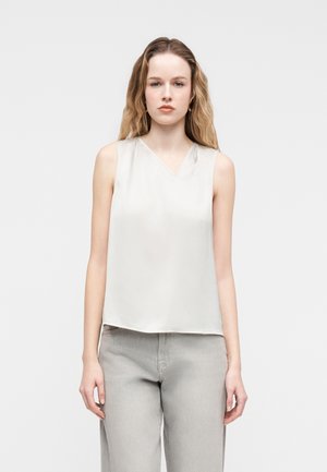 Γυναίκα με μακριά κυματιστά μαλλιά φοράει αμάνικο λευκό πουκάμισο και ανοιχτό γκρι παντελόνι, στέκεται μπροστά σε απλό ανοιχτό γκρι φόντο.