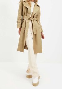 Žena v béžovém trenčkotě svázaném v pase, krémových kalhotách a bílých platformových teniskách, stojící na bílém pozadí.