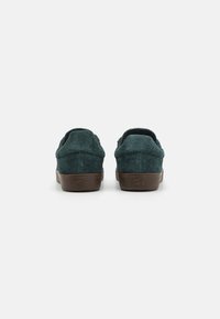 Zapatos de ante verde tipo slip-on con suela de goma marrón, que presentan una puntera redonda y una textura suave. Vista trasera, sin cordones ni acentos visibles.