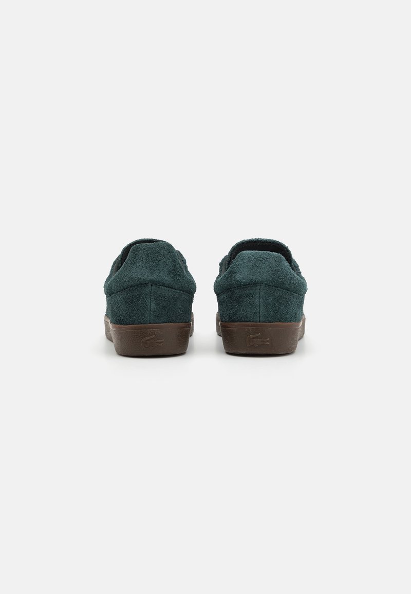 Zapatos de ante verde tipo slip-on con suela de goma marrón, que presentan una puntera redonda y una textura suave. Vista trasera, sin cordones ni acentos visibles.