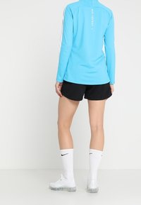 Camiseta atlética de manga larga en azul claro con cuello simulado, combinada con pantalones cortos negros, calcetines blancos hasta la rodilla y zapatillas transparentes, con acentos de logo.