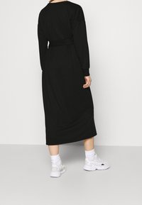 Musta pitkähihaisen mekko vyöllä, joka on valmistettu pehmeästä kankaasta. Siinä on suora leikkaus, polven pituinen helma, ja se on yhdistetty valkoisiin urheilukenkiin ja sukkiin.
