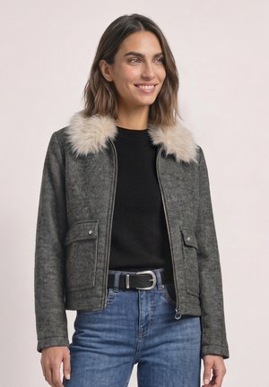 Femme portant une veste grise zippée avec un col en fourrure claire, un haut noir, un jean bleu et une ceinture noire, souriante et regardant vers la droite.