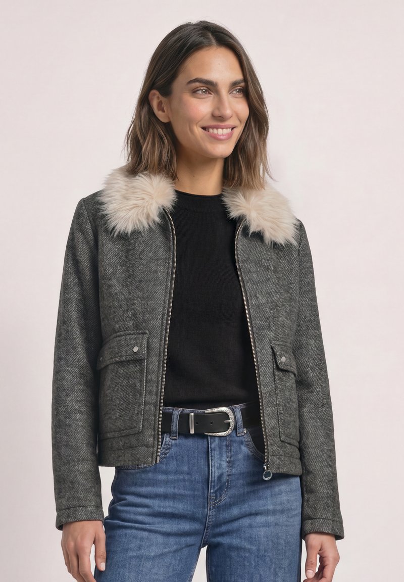 Femme portant une veste grise zippée avec un col en fourrure claire, un haut noir, un jean bleu et une ceinture noire, souriante et regardant vers la droite.