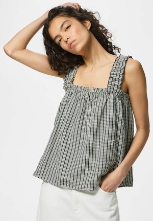 Frau mit lockigem Haar trägt ein ärmelloses schwarz-weiß kariertes Top und weiße Hose, posiert mit einer Hand in der Tasche und der anderen hinter dem Kopf.