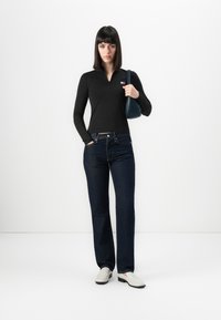 Vrouw in een zwart longsleeve top, donkerblauwe spijkerbroek, witte schoenen, met een marineblauwe handtas, staande tegen een witte achtergrond.