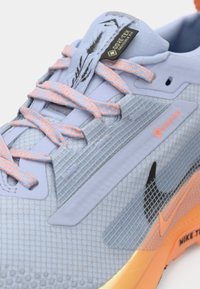 Pantofii sport de culoare albastru deschis, cu partea superioară din plasă, accente portocalii, suprapunerile texturate și brandul Gore-Tex. Au șireturi reflectorizante și logo negru.