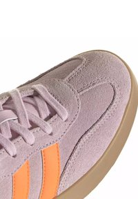 Gros plan sur la pointe d'une basket en daim mauve avec des lacets roses, des bandes latérales orange et une semelle en caoutchouc beige sur fond blanc.