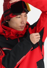 Rot schwarze Skijacke mit einem Reißverschluss unter dem Arm, verstellbarer Kapuze und reflektierenden Akzenten. Begleitet von einer roten und schwarzen Mütze und einer Skibrille.