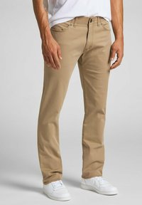 I pantaloni di cotone beige presentano un design a gamba dritta, cinque tasche e accessori in metallo, abbinati a sneakers bianche per un contrasto elegante.