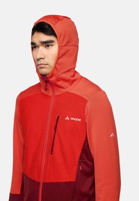 Rote Kapuzenjacke mit strukturiertem oberen Teil, glattem unteren Abschnitt und kontrastierenden dunkleren roten Akzenten. Verfügt über einen Reißverschluss und ein Logo auf der Brust.