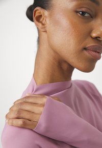 Langermet topp med høy hals i lys lavendel-fabric, med en glatt tekstur og tettsittende design, med en hånd som hviler på skulderen.
