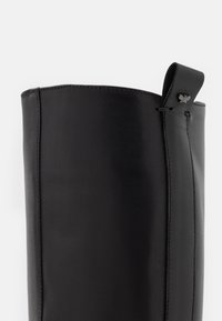 Botte noire en cuir montant jusqu'au genou avec une texture lisse, un bord supérieur arrondi, et une boucle en cuir décorative avec un détail en métal à l'arrière.