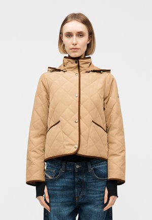 Kvinde iført en beige vatteret jakke med brune kanter over blå jeans, stående mod en ensfarvet lys baggrund.