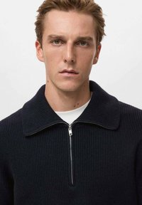 Maglione blu navy a costine con zip frontale e ampio colletto. Presenta un tessuto a maglia testurizzato e una maglietta bianca visibile sotto.