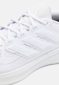 Chaussure de sport blanche avec une tige en tissu texturé, trois rayures blanches, lacets plats, et une semelle en caoutchouc avec un motif de traction.