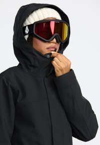 Schwarze wasserdichte Jacke mit Kapuze, ausgestattet mit hohem Kragen und Reißverschluss. Kombiniert mit übergroßen Skibrillen mit getönten Gläsern.