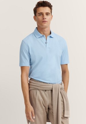 Helles blaues Polohemd mit Kragen und zwei Knöpfen, kombiniert mit beigen Hosen und einem hellbraunen Pullover, der um die Taille gebunden ist.