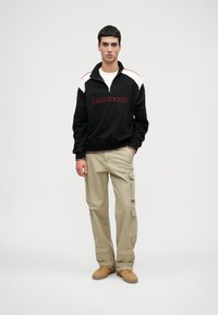 Μαύρη φούτερ μπλούζα με φερμουάρ και λευκές και κόκκινες λεπτομέρειες; γραφικό "LET'S FRANS". Μπεζ cargo παντελόνι με πλαϊνές τσέπες. Καφέ παπούτσια. Απλό φόντο.