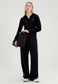 Schwarze Jacke mit Reißverschluss, langen Ärmeln und tailliertem Schnitt, kombiniert mit losen schwarzen Hosen mit Seitenstreifen. Eine schwarze Tasche wird in der Hand gehalten.