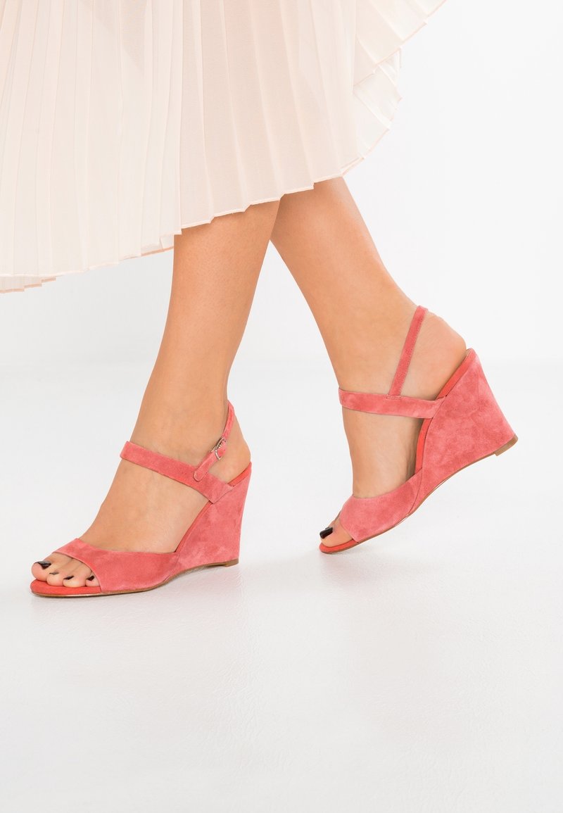 Femme portant des sandales compensées en suède corail rose avec des brides à la cheville et une jupe blanche plissée, debout sur un sol blanc.