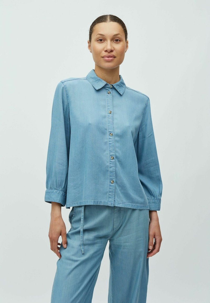 Chemise en denim bleu clair avec un col à revers, une fermeture à boutons et des manches longues. Le tissu présente une texture lisse et une coupe décontractée.