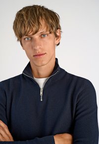 Marinefarbener Pullover mit Reißverschluss und strukturiertem Stoff. Verfügt über einen silbernen Reißverschluss, eine weiße innere Schicht und ein tailliertes Design, gezeigt vor einem schlichten Hintergrund.