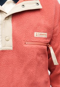 Ζακέτα από φλις Columbia σε κοραλί ροζ χρώμα με μπεζ κουμπιά τύπου snap, περιμετρική διακόσμηση στο γιακά και τσέπη στο στήθος με φερμουάρ στο πάνω μέρος του κορμού.