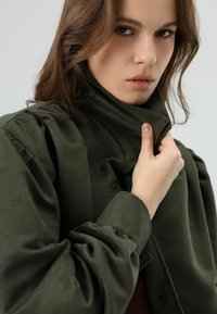 Veste vert olive avec un col montant, des manches bouffantes et un tissu texturé. Gros plan sur une main saisissant le col, mettant en valeur les détails des boutons.