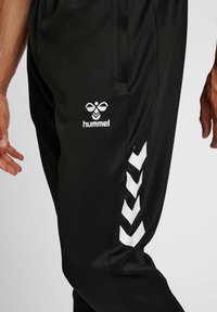 Close-up of black sports pants with white Hummel logo and chevron stripes on a person's leg against a plain background.