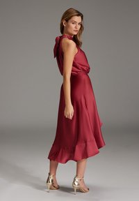 Femme portant une robe en satin bourgogne sans manche avec un nœud à l'arrière, chaussée de sandales à talons dorés, se tenant sur un fond gris neutre.