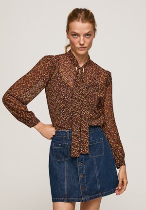 Blusa floreale marrone con maniche lunghe e dettaglio a fiocco al collo. Abbinata a una gonna in denim blu con tasche frontali e chiusura a bottoni.