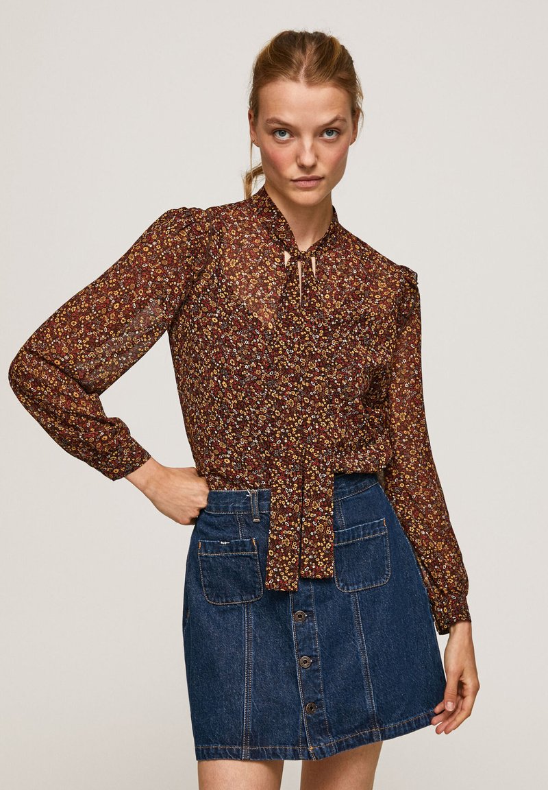 Blusa floreale marrone con maniche lunghe e dettaglio a fiocco al collo. Abbinata a una gonna in denim blu con tasche frontali e chiusura a bottoni.