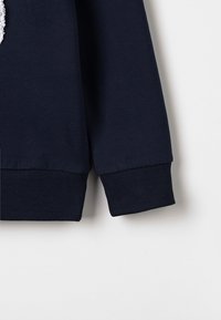 Manica di felpa blu marina con polsino a coste, che presenta un accento testurizzato e un tessuto liscio, mostrando un contrasto di materiali e finiture.