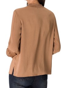 Blouse à manches longues, texturée, de couleur marron clair, avec un col froncé, des fentes latérales et une coupe décontractée, associée à un jean en denim foncé.
