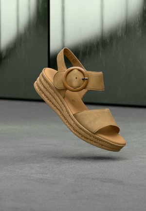 Beige wedge sandal with soft suede straps, open toe, circular wooden accent, and textured rubber sole for grip.