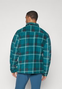 Muž, ktorý má na sebe svetlomodro a tmavomodro károvanú fleecovú bundu, otočený chrbtom, v páre s modrými nohavicami pred jednoduchým svetlým pozadím.