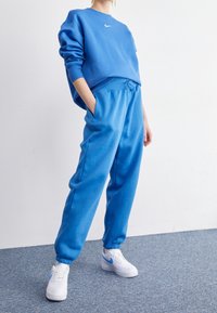 Person som bär en blå Nike-tröja och matchande sweatpants med vita Nike-sneakers, står på en grå matta mot en vit vägg.