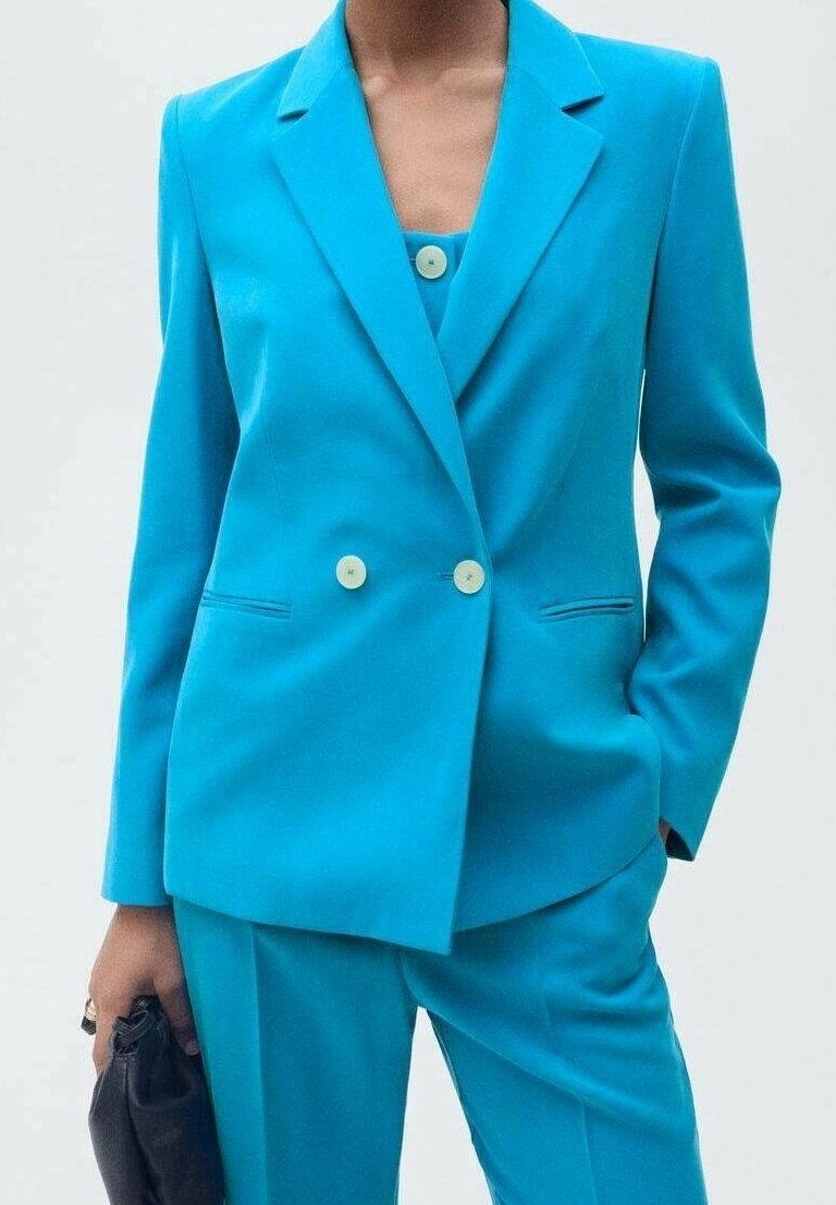 Femme portant un costume bleu vif à double boutonnage avec des boutons blancs, tenant une petite pochette noire dans sa main gauche.