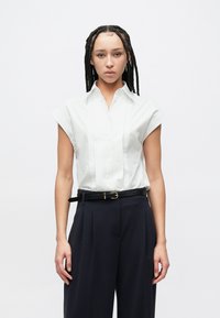 Άσπρο πουκάμισο με κοντά μανίκια, λαιμόκοψη με γιακά και πιέτες από μπροστά, συνδυασμένο με ψηλόμεσο σκούρο μπλε παντελόνι και μαύρη ζώνη.