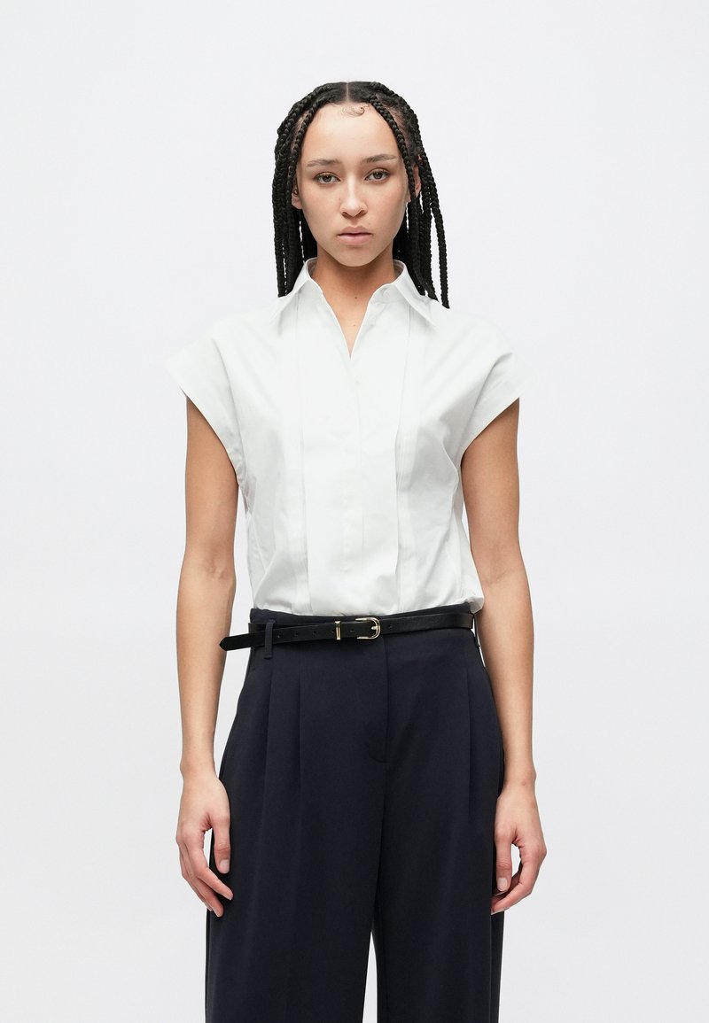 Άσπρο πουκάμισο με κοντά μανίκια, λαιμόκοψη με γιακά και πιέτες από μπροστά, συνδυασμένο με ψηλόμεσο σκούρο μπλε παντελόνι και μαύρη ζώνη.