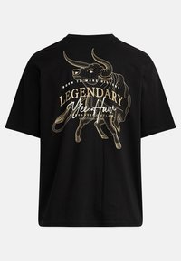 Črna majica s kratkimi rokavi, značilna z zlatim grafičnim motivom bika in napisom "Legendary Yee Haw" na hrbtni strani. Ohlapen kroj, bombažni material.