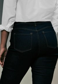 Los jeans de mezclilla oscura presentan un diseño ajustado con dos bolsillos traseros y sutiles detalles de costura, combinados con una camisa blanca cerca de la cintura.