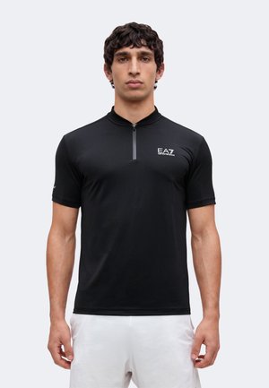 Bărbat care poartă un tricou negru cu mâneci scurte cu fermoar și logo EA7 Emporio Armani, stând în fața unui fundal simplu și luminos.